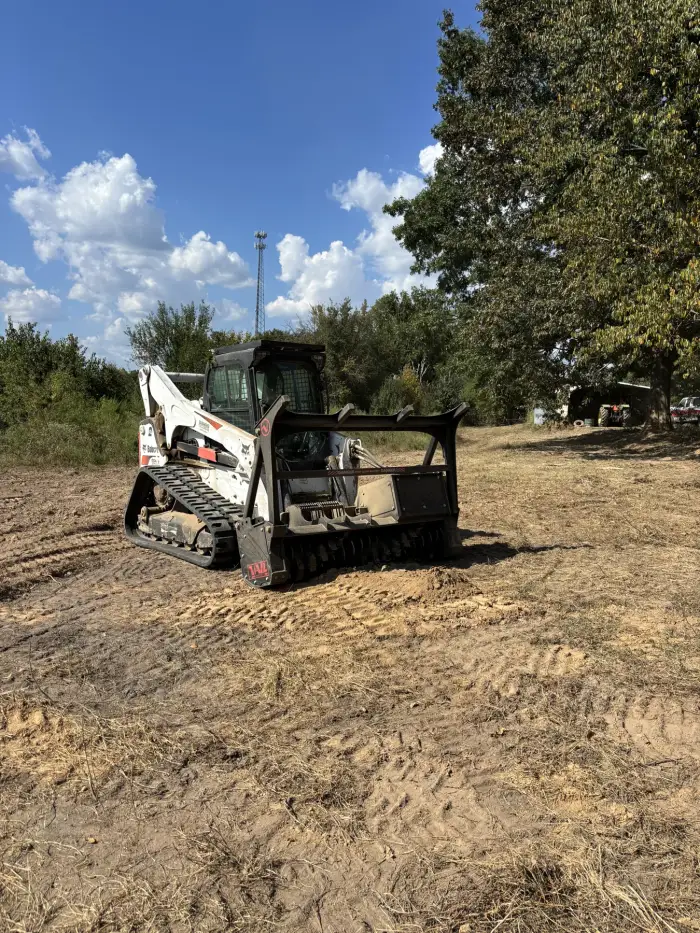 A compact track loader fitted with a forestry mulching head standing on freshly mulched ground under a blue sky